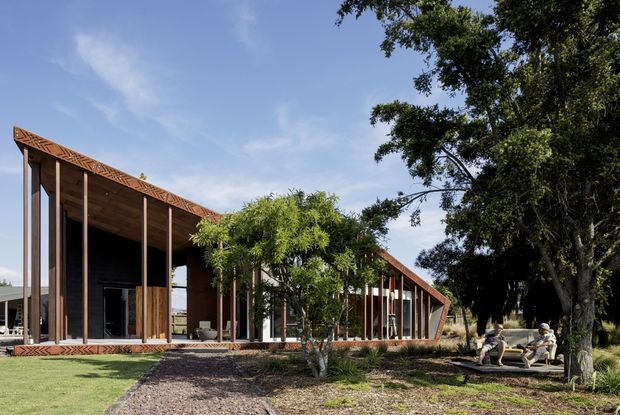 A low, elongated contemporary pavilion with a strongly expressed pitched roof and exposed timber truss detailing. Floor-to-ceiling glazing and slender timber posts open the building toward a landscaped lawn and shaded garden, with a gravel path leading to the entrance. Mature trees and informal outdoor seating frame the scene.