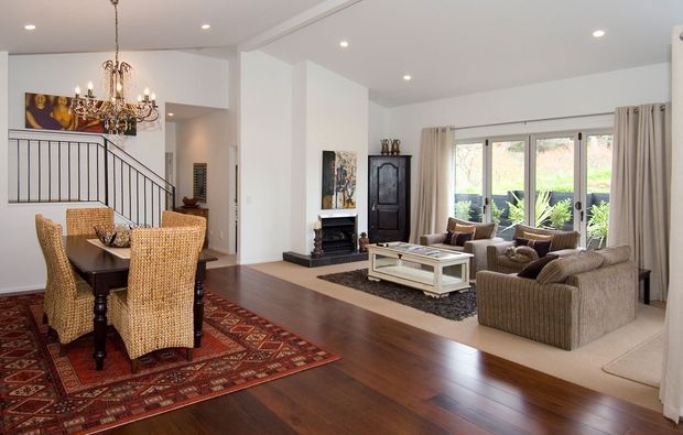 Open-plan formal dining and living area with dark timber flooring, a patterned area rug, woven dining chairs around a dark table, upholstered lounge seating, a glass-topped coffee table, a traditional fireplace surround, ornate chandelier, staircase with black balustrade, and large windows dressed with neutral curtains.