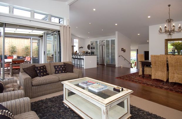 Open-plan residential interior with a living room in the foreground, dining area to the right, and kitchen beyond. The room features a large striped sectional sofa, distressed white coffee table, dark rug, polished dark timber flooring, white walls, extensive glazed doors and windows, and a traditional chandelier over a dark wood dining table with woven dining chairs. The layout connects to a bright glazed outdoor space or courtyard.
