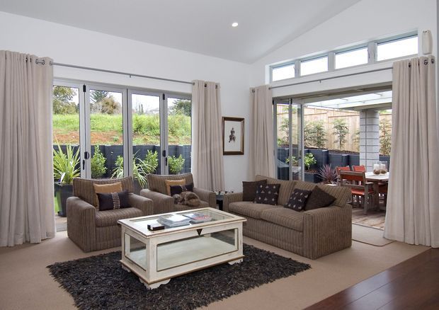 Bright, spacious living room with large glazed doors and high clerestory windows opening toward a landscaped outdoor dining area. The room features two brown upholstered sofas, a glass-topped coffee table with a light painted frame, neutral curtains, beige carpet, and a dark textured rug. The interior has a relaxed, refined contemporary-traditional feel with abundant natural light.