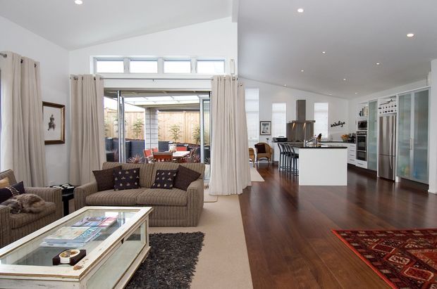 Bright open-plan living, dining and kitchen interior with white walls, recessed ceiling lights, dark timber flooring, and large glazed doors opening to an outdoor area. The living zone has upholstered sofas, a glass-topped coffee table and layered rugs, while the kitchen features a white island with seating, dark benchtops, extensive cabinetry and stainless-steel appliances. Neutral curtains and furnishings create a relaxed, refined residential setting.