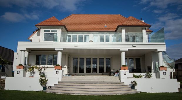Large multi-storey suburban residence viewed from the front, with a symmetrical white rendered facade, broad upper and lower terraces, classical-style columns and balustrades, extensive glazing, and a terracotta tiled hipped roof. Wide steps lead up from the lawn to the main entrance terrace.