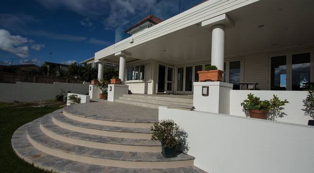 Single-storey residential exterior with a broad covered front terrace, large round columns, white horizontal cladding, glazed doors and windows, and a curved flight of tiled steps leading up from the lawn. Potted plants and a low white boundary wall frame the entry, with a flat roof and rooftop structure visible above.