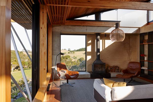 A warm, light-filled contemporary living room with expansive glazing overlooking a rural landscape. The room features exposed timber ceiling slats and beams, timber-framed windows, textured earthen-toned walls, a freestanding wood-burning stove, built-in shelving, leather lounge chairs, and a large upholstered sofa. A sculptural woven pendant adds a natural decorative element.