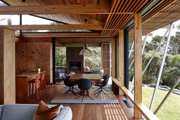 A warm, timber-lined open-plan living and dining space with a compact built-in bar at left, a dining table and sculptural chairs in the centre, and a fireplace lounge beyond. Extensive glazing and open sides connect the room to a landscaped garden, while timber pergola slats cast strong linear shadows across the floor. The space has a refined contemporary pavilion-like design with exposed timber beams and posts.