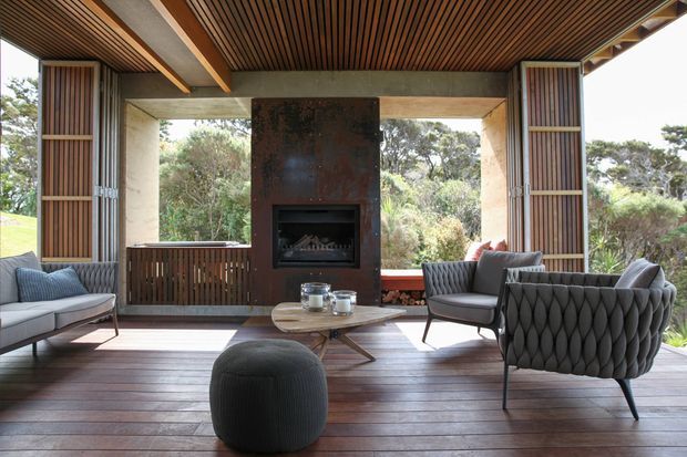 A refined, open-sided living room or lounge with expansive glazing overlooking dense natural bushland. The space features a dark timber slatted ceiling, wide timber floorboards, exposed concrete beams and wall surfaces, vertical timber screens, and a central sculptural fireplace clad in dark oxidised metal. A grey sofa, quilted lounge chairs, round ottoman, and low timber coffee table create a warm, sophisticated seating area.