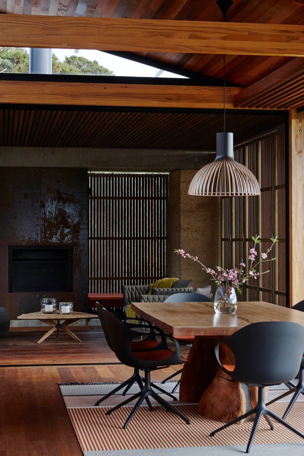 A dramatic, warm-toned open-plan living and dining interior with a strong timber structure and pitched ceiling. The room features a substantial timber dining table, sculptural dark dining chairs, a modern suspended pendant light, a low lounge area with sofa and coffee table, a dark fireplace wall, vertical timber screening, and expansive timber flooring. Natural light enters through high-level glazing and screened openings, creating a sophisticated, tactile atmosphere.