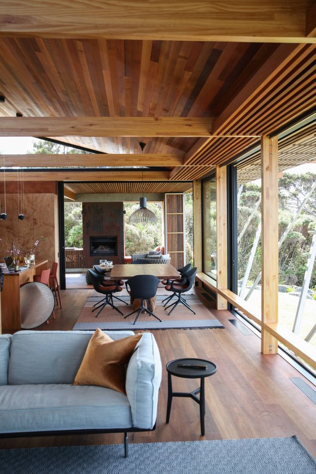 A warm, open-plan timber-lined interior combining a living room and dining area. The space features extensive floor-to-ceiling glazing, exposed timber beams, a slatted timber ceiling, timber flooring, a dining table with contemporary chairs, a grey sofa, a built-in fireplace, and strong visual connections to surrounding vegetation.