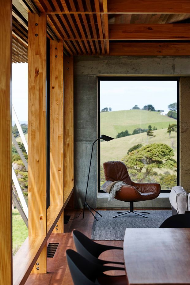 A warm, timber-framed interior overlooking rolling green hills. Exposed timber ceiling beams and slatted rafters contrast with a raw concrete wall and large black-framed window. The space includes a brown leather lounge chair, black floor lamp, dining table and chairs, timber flooring, and a textured grey rug, creating a refined modern retreat.