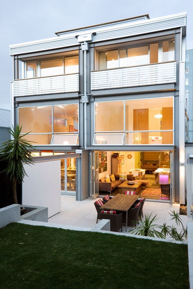 Exterior view of a contemporary multi-storey urban residence with a silver-grey metal facade, extensive glazing, projecting balconies with horizontal slatted balustrades, and a roof-level terrace railing. The lower level opens onto a paved courtyard and outdoor dining area, with a warmly lit living room visible through the glazed doors. The foreground includes a small lawn, built-in concrete planter structures, and tropical-style planting.
