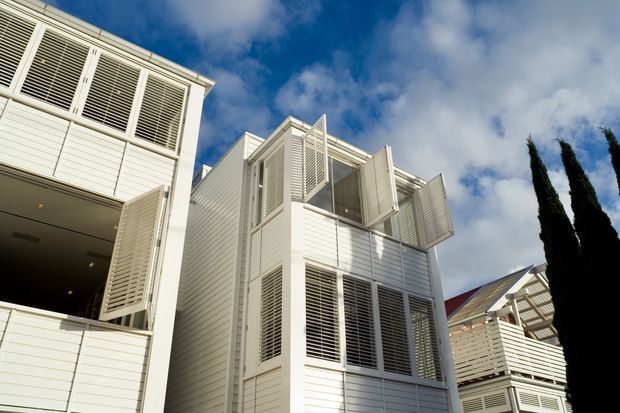 Upward-looking exterior view of a contemporary multi-storey residential building with white horizontal weatherboard cladding, extensive louvred and screened windows, projecting balconies, and open architectural ventilation panels. A neighbouring structure and tall narrow trees are visible at the edge, beneath a partly cloudy blue sky.