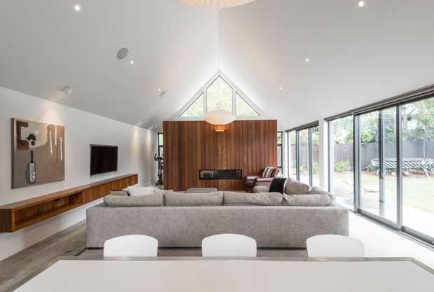 Bright contemporary living room with a dramatic vaulted ceiling, white walls, polished concrete flooring, and extensive floor-to-ceiling glazing along one side. A large light-grey sectional sofa faces a timber feature wall with an integrated fireplace and television, while a dining table occupies the foreground. The triangular clerestory window and exposed timber wall create a strong architectural focal point.