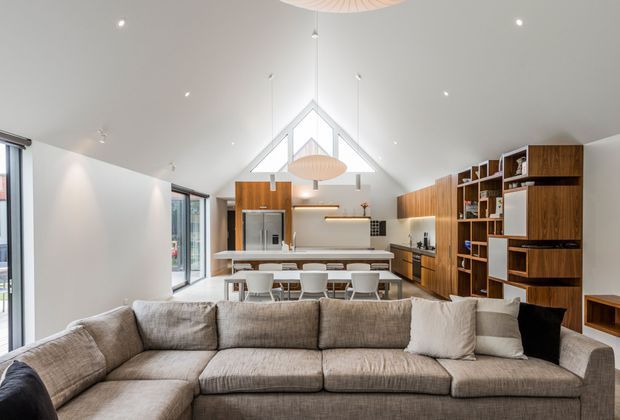 A bright open-plan living, dining and kitchen interior viewed from behind a large sectional sofa. The space features a high vaulted ceiling with exposed triangular glazing and pendant lights, extensive white walls, warm timber kitchen cabinetry and built-in shelving, a long island with dining stools, integrated appliances, and large glazed openings along the left side. The styling is clean, contemporary and warm with a neutral palette.