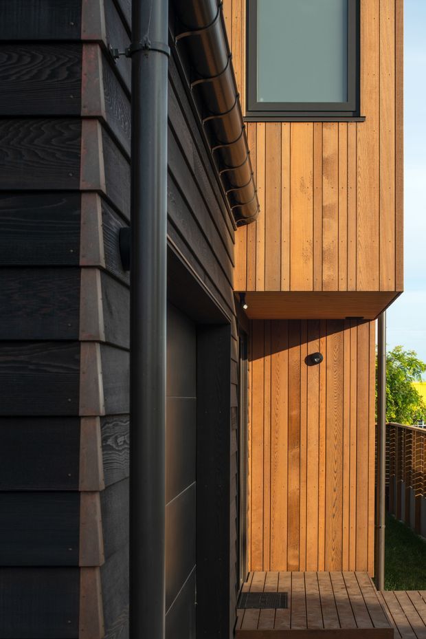 Close-up architectural exterior detail of a contemporary house, showing contrasting dark horizontal cladding and warm vertical timber weatherboards, black-framed window and door elements, a metal gutter and downpipe, and a small timber deck or landing. The image focuses on facade materials, junctions, and projecting upper-storey form.