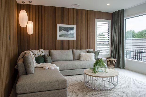 A refined living room with a large L-shaped grey sectional sofa, round wire-frame coffee table, textured rug, timber-lined feature wall, sculptural pendant lights, and full-height glazing overlooking a balcony or exterior view. The room has a warm, contemporary feel with layered neutral textiles and dark curtains.