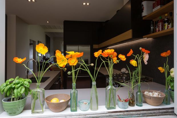 A dark, contemporary kitchen viewed through a serving window or opening, with black cabinetry, warm timber shelving, integrated under-shelf lighting, a cooktop, sink and island bench. Several glass vases of bright orange flowers and potted greenery line the foreground ledge, adding vivid colour to the otherwise restrained interior.