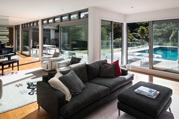 Bright contemporary living room with a large charcoal sectional sofa and matching ottoman, light timber flooring, and extensive floor-to-ceiling glazing. The glazing opens to a pool terrace and landscaped garden, while a dining area and additional seating are visible within the open-plan interior. Clerestory windows and slim dark frames provide abundant natural light.