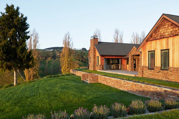 Exterior view of a large rustic lodge-style residence set on a landscaped hillside. The building features warm timber cladding, shingle detailing in the gable, substantial natural stone masonry, dark pitched roofs, broad glazing, and a raised stone terrace. Mature trees, lawn, and rolling wooded hills surround the property.