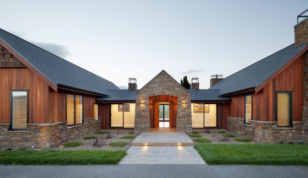 Symmetrical single-storey residential building with a central stone entry pavilion and wings extending to both sides. The facade combines dark grey pitched roofs, vertical warm timber cladding, and low rough-stone walls, with glazed doors and windows arranged around a landscaped courtyard approach.