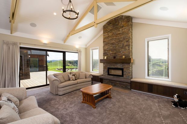 A bright, spacious living room with a vaulted ceiling and exposed pale timber beams. The room features a large stacked-stone fireplace with a timber mantel, two upholstered sofas, a timber coffee table, broad carpeted flooring, and extensive sliding glass doors opening to a deck and landscape beyond. Neutral furnishings, curtains, and window blinds create a warm, relaxed interior.