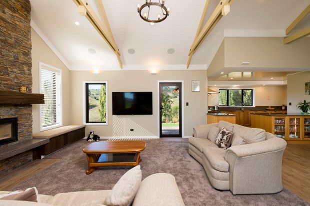 A warm, spacious open-plan living room with a large stone fireplace, built-in timber hearth cabinetry, television, plush sectional seating, coffee table, and carpeted floor. Exposed decorative timber ceiling beams and a circular pendant light add architectural character. The kitchen and dining area are visible beyond the living space, with timber cabinetry and a window overlooking greenery.
