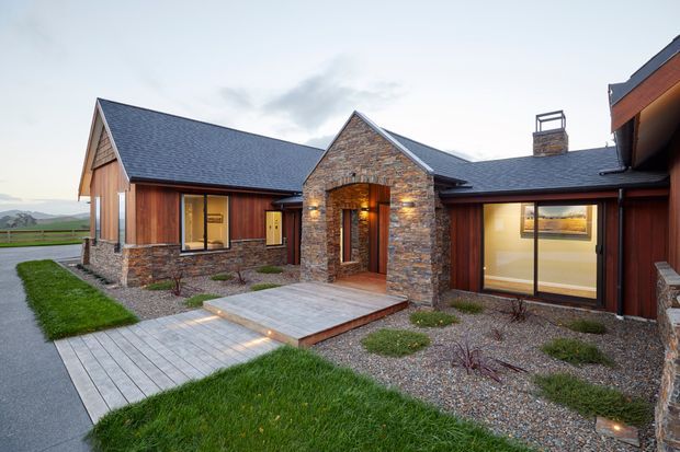 Single-storey contemporary lodge-style residence viewed from the landscaped front approach. The building has several connected gabled volumes, dark grey pitched roofs, extensive natural stacked-stone masonry, warm timber cladding, large glazed openings, and a stone-framed recessed entry. A timber pedestrian deck and gravel garden beds lead to the entrance, with open rural landscape visible beyond.