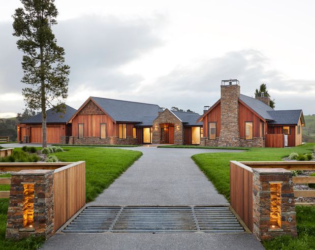 Exterior view of a sprawling single-storey lodge-style residence arranged around a central entry courtyard. The building features timber-clad gabled wings, dark pitched roofs, prominent natural-stone chimney and entry detailing, warm recessed lighting, manicured lawns, and a paved approach crossing a metal cattle grid. A rural landscape is visible beyond the property.