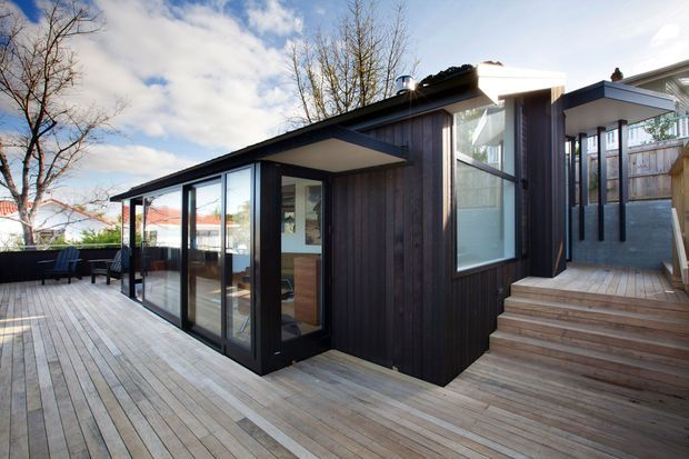 A contemporary single-storey extension or pavilion viewed from a timber deck. The building has dark vertical timber cladding, extensive black-framed floor-to-ceiling glazing, a low-pitched roof with broad overhangs, and timber steps leading to a raised entrance. A light-filled interior is visible through the glazing.