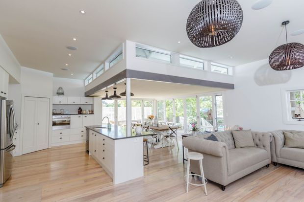 Bright open-plan kitchen, dining, and living area with a white island, light timber flooring, extensive glazing, and a raised mezzanine-like upper level overlooking the room. The space features woven pendant lights, white cabinetry, a pale sectional sofa, and a high clerestory band that brings in abundant daylight.