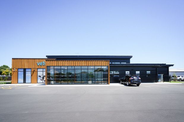 A contemporary low-rise commercial building viewed from across a broad paved forecourt. The facade combines dark horizontal cladding with warm vertical timber-look slats, extensive floor-to-ceiling glazing, and a flat roofline. A blue 'vet' sign and parked vehicles indicate a veterinary practice or similar commercial facility.