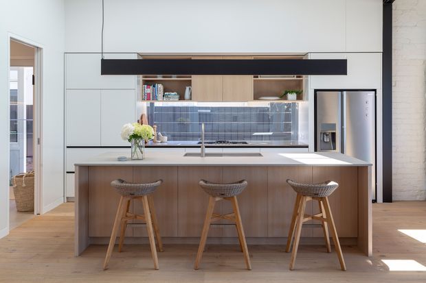 Contemporary open-plan kitchen with a large timber island, three woven-seat bar stools, pale stone benchtops, integrated cabinetry, dark blue-grey tiled splashback, and a prominent black suspended rangehood. A stainless-steel refrigerator, open shelving, timber flooring, and exposed white-painted masonry are also visible.