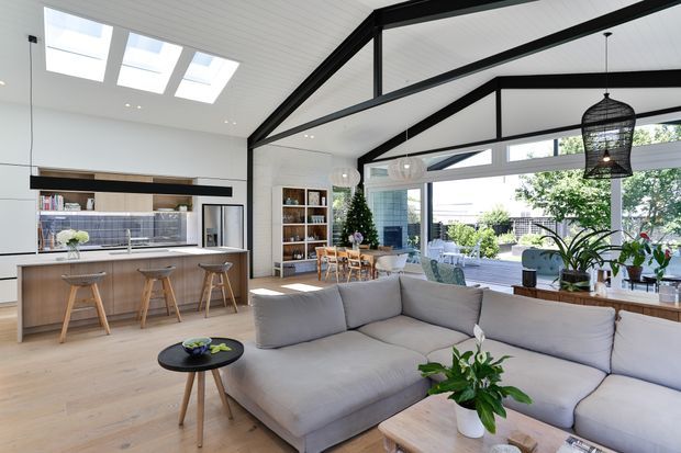 Bright open-plan living and kitchen interior with a large light-grey sectional sofa in the foreground, timber flooring, a timber kitchen island with stools, open shelving, dining area, extensive glazing, skylights, and a dramatic black exposed roof structure. Indoor plants and woven pendant lights add a natural contemporary character.