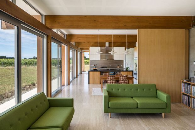 A bright, open-plan contemporary living and kitchen interior with a long wall of floor-to-ceiling glazing overlooking a rural landscape. Green mid-century-style sofas sit on a pale timber floor, while exposed warm timber beams span the ceiling and frame the space. The kitchen features a central island, pale cabinetry, pendant lighting, and timber or wood-toned wall joinery. A white tiled or plasterboard ceiling and clean linear detailing create an airy, refined atmosphere.