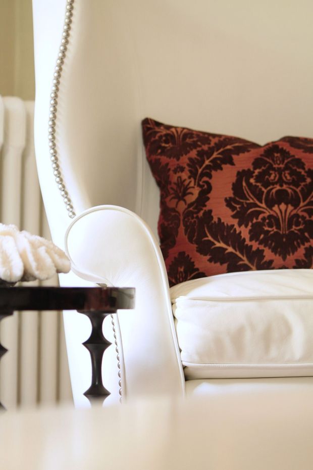 Close-up interior detail of an elegant upholstered white armchair with rounded contours and decorative nailhead trim, paired with a dark wood side table. A rich burgundy and dark brown damask cushion provides contrast against the warm cream wall and soft neutral furnishings.