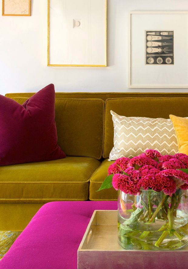 Close-up interior view of a richly coloured living room, centred on a mustard olive velvet sofa with burgundy, cream patterned and yellow cushions. A vivid magenta upholstered ottoman and wooden tray table sit in the foreground, topped by a glass vase of bright pink flowers. White walls display framed artwork with gold-toned trim.
