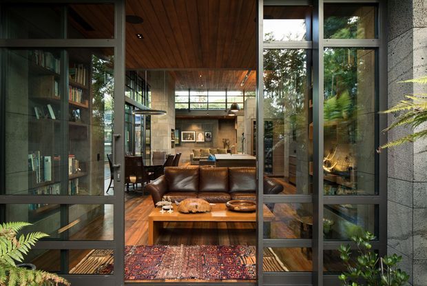 View through open-framed glazed doors into a sophisticated open-plan living space. The room features a large brown leather sofa, timber coffee table, patterned rug, dark timber flooring and ceiling, extensive steel-framed glazing, built-in shelving, and a kitchen/dining area beyond. Concrete or masonry walls and abundant indoor planting create a warm, refined indoor-outdoor atmosphere.