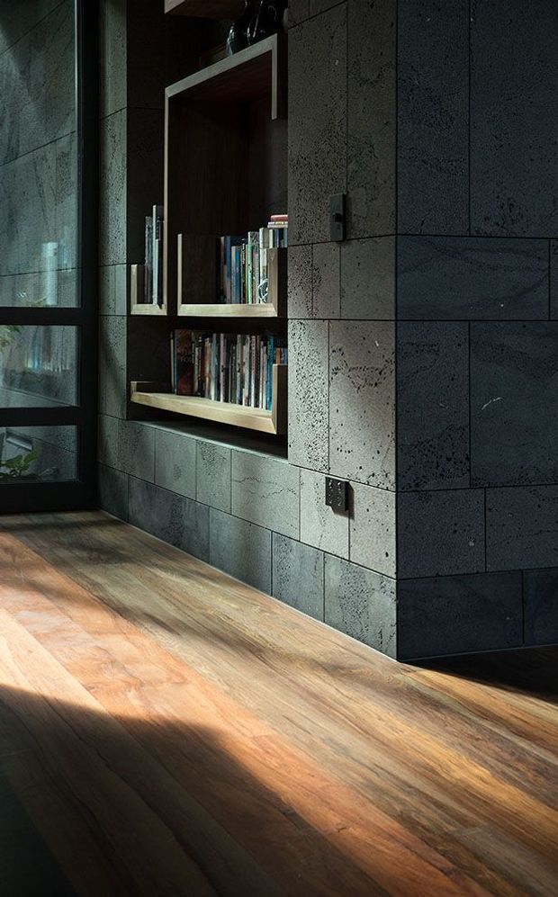 A moody interior detail showing a dark stone or concrete-block feature wall with a recessed built-in timber bookshelf filled with books. A tall black-framed glazed door or window is visible at left, while warm timber flooring catches strong sunlight across the foreground.