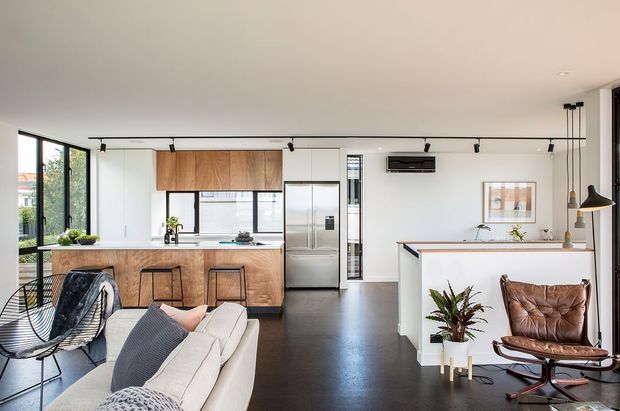 Open-plan contemporary interior combining a kitchen, living area, and dining space. The kitchen features warm timber cabinetry, a central island with stools, stainless-steel appliances, and dark flooring. A white sofa, wire lounge chair, and leather lounge chair furnish the living area, while black track lighting, large glazed doors, and minimal white walls create a bright modern setting.
