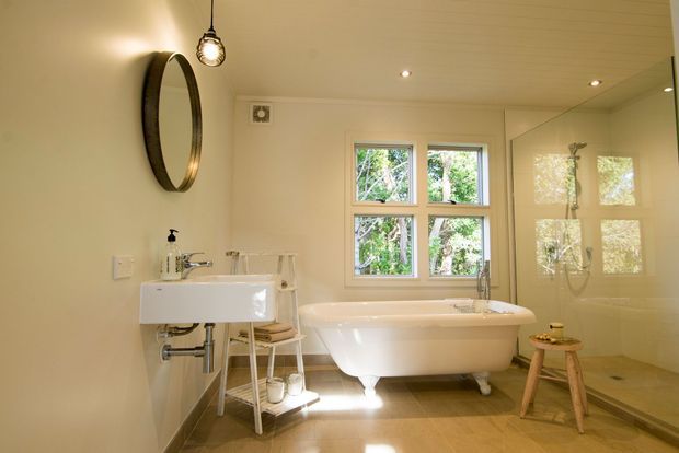 Bright, warm-toned bathroom featuring a freestanding white clawfoot-style bathtub beneath a four-pane window, a wall-mounted white basin with exposed plumbing, a frameless glass shower enclosure, and a round wall mirror. Timber-look flooring, cream walls, and pendant and recessed lighting create a relaxed contemporary-traditional feel.