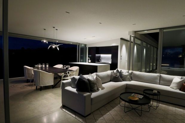 A sophisticated open-plan living, dining and kitchen area at night, featuring a large light-grey sectional sofa, dark round coffee tables, a dark dining table with upholstered chairs, polished concrete flooring, extensive floor-to-ceiling glazing, and a minimalist kitchen island with illuminated cabinetry. Contemporary pendant lighting and the dark exterior outlook create a refined, moody atmosphere.