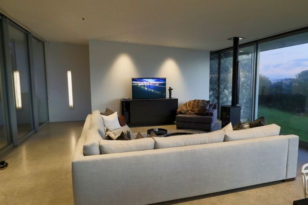 Contemporary living room with a large light upholstered sectional sofa arranged around a low coffee table and dark media console. Floor-to-ceiling glazing on the right provides views of the landscaped exterior, while a freestanding black wood-burning stove stands near the windows. The room has smooth pale walls, a minimalist ceiling, polished concrete flooring, and dark-framed sliding glass doors along the left.