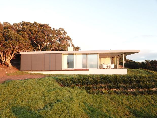 Single-storey modern house set in a grassy, gently sloping landscape, with a long low rectangular form, broad flat roof overhang, extensive floor-to-ceiling glazing, and a covered outdoor living area. The facade combines warm brown vertical cladding panels with pale rendered walls and dark-framed glazing, backed by mature trees.