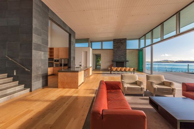 Open-plan contemporary living space with a kitchen, dining area and lounge arranged beside expansive floor-to-ceiling glazing. Warm timber flooring and ceiling boards contrast with dark stone-clad walls, a central fireplace, and a vivid red sectional sofa. The glazing overlooks a swimming pool and distant waterfront landscape, while a stairway leads to an upper level at left.