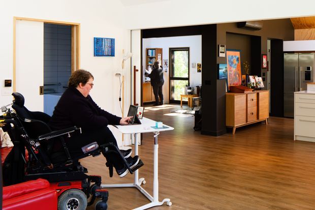 An accessible open-plan residential interior with warm timber flooring, white walls, and black architectural partitions. The foreground contains a wheelchair user at a compact white work table, while the background includes a timber sideboard, artwork, shelving, a doorway, and part of a contemporary kitchen with stainless-steel refrigeration and white cabinetry. The space appears bright, functional, and designed for accessible living.