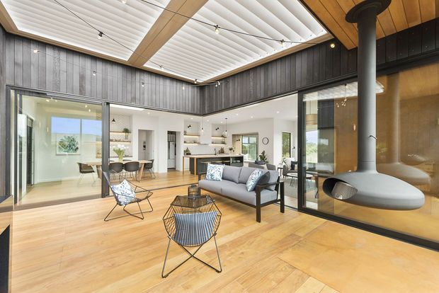 A bright, open-plan contemporary living space with warm timber flooring and ceiling lining, dark vertical wall cladding, extensive glazing, and a prominent suspended wood-burning fireplace. The room opens directly into a modern kitchen and dining area, with mid-century-inspired lounge furniture and a large clerestory-style roof opening fitted with white louvres.