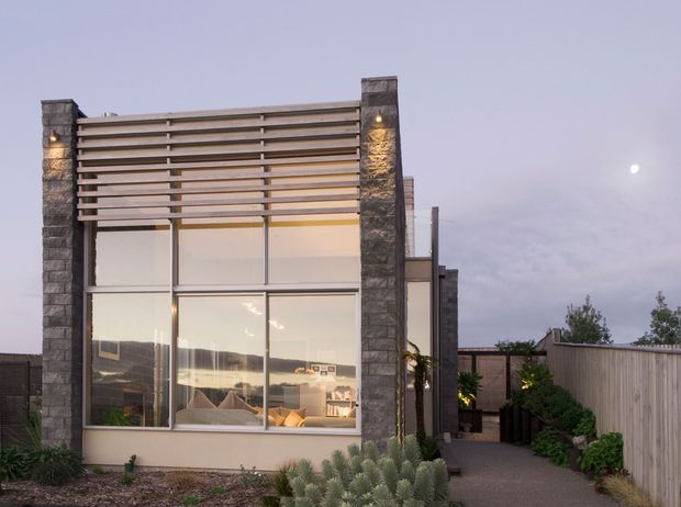 Contemporary single-storey residential facade at dusk, featuring extensive floor-to-ceiling glazing, tall dark stacked-stone piers, horizontal timber screening above the windows, and warm integrated lighting. A landscaped path and timber boundary fence frame the approach, while a furnished living area is visible through the glass.