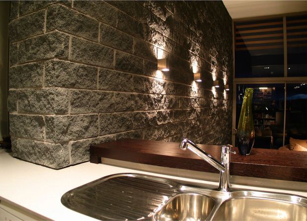 A warmly lit kitchen or bar servery with a stainless-steel double sink and chrome tap in the foreground. A dark timber benchtop runs along a dramatic rough stone-block feature wall, accented by evenly spaced wall lights. A large mirror or glazed opening reflects shelving and the room beyond.