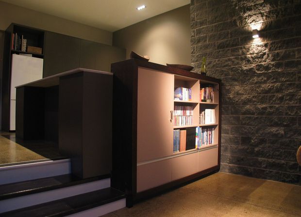 Dimly lit contemporary interior featuring dark built-in cabinetry and a partially open bookcase with integrated lighting, set against a textured dark masonry feature wall. A short flight of steps leads toward a compact kitchen or utility area, with polished concrete flooring and recessed ceiling lighting.