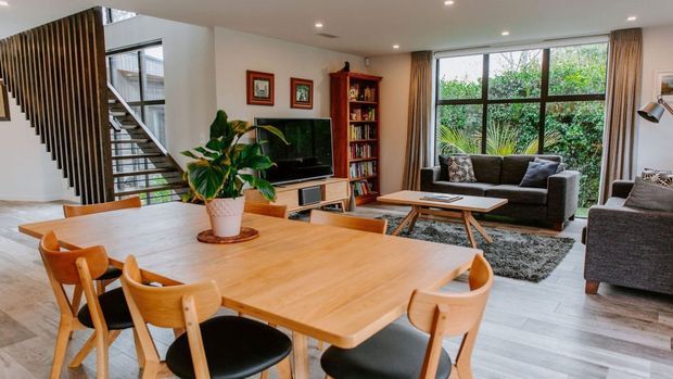 A bright open-plan living and dining area with a large timber dining table and wooden chairs in the foreground, a grey sofa seating area with a television and built-in timber shelving, and a modern staircase with vertical timber slats. Large windows overlook greenery, while neutral finishes and warm timber furnishings create an inviting contemporary interior.