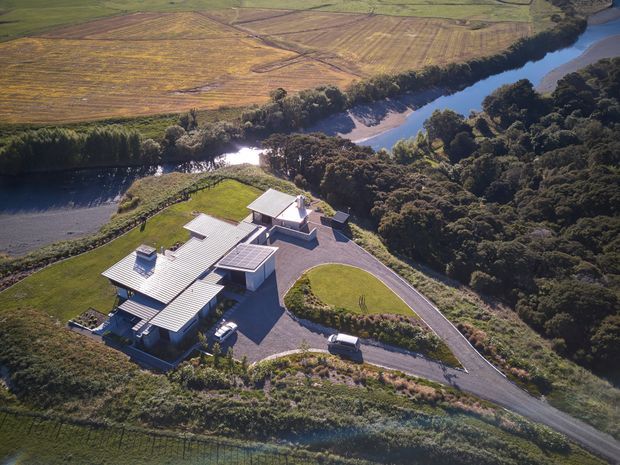 Aerial view of a contemporary multi-wing building set on a landscaped, elevated site beside a winding river. The property has multiple low-pitched corrugated metal roof forms, light-coloured walls, paved access drives, lawns, and dense surrounding vegetation, with agricultural fields visible across the water.